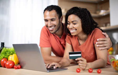 Cheerful young african american wife and husband at table with laptop, credit card and organic...