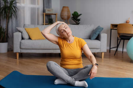 Healthy lifestyle concept. Senior woman stretching neck, sitting on yoga mat and warming up before domestic workoutの写真素材