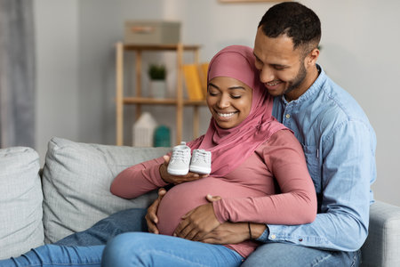 Happy Black Muslim Spouses Holding Small Baby Shoes While Relaxing On Couch