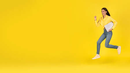 Happy Woman Running Holding Laptop Looking Aside Over Yellow Backgroundの写真素材