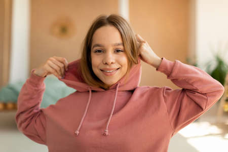 Portrait of positive teen girl wearing hood and smiling at camera, having fun and resting in bedroom interior at homeの写真素材