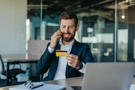 Mobile payments. Happy male worker holding credit card and talking on cellphone with customer...
