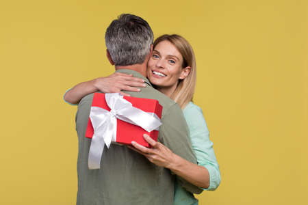 Happy middle aged spouses hugging on yellow background, woman holding gift box and smiling at cameraの写真素材