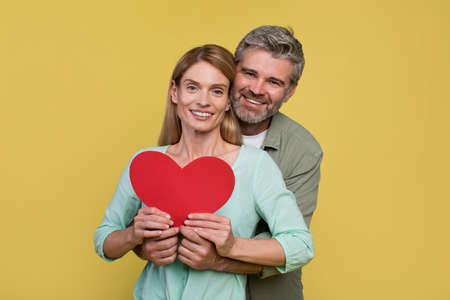 Loving middle aged spouses holding paper heart, man embracing wife brom back and smiling together at cameraの写真素材