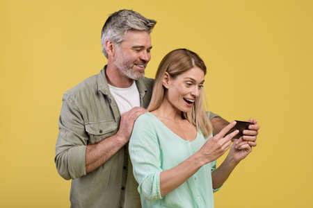 Excited middle aged spouses looking at cellphone screen, using new application, yellow studio...