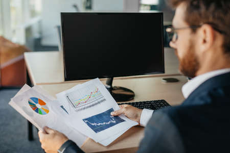 Successful entrepreneur reading financial reports, sitting at workplace with computer with black...