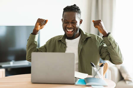 Overjoyed black man gesturing YES in front of laptop, celebrating success, sitting at table at home officeの写真素材