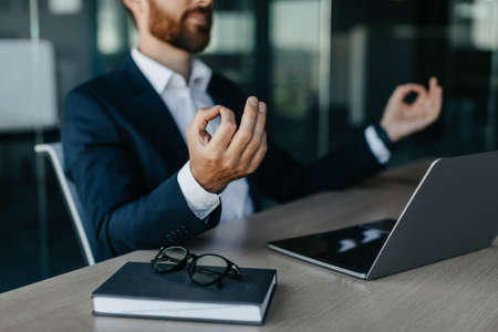 Relaxed businessman meditating at workplace in office, calm male entrepreneur in suit sitting at desk, selective focusの写真素材