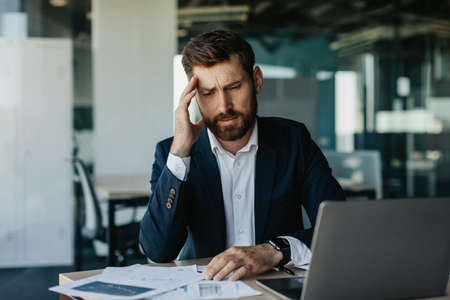 Stressed entrepreneur suffering headache, sitting at desk with lot of papers, tired businessman having acute migraineの写真素材