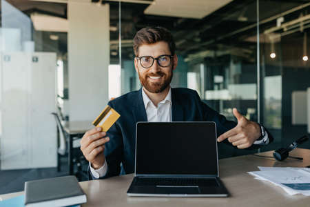 Happy businessman holding credit card and pointing at laptop screen with empty screen, sitting in office, mockupの写真素材