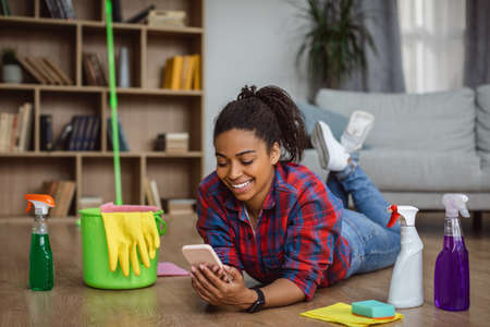 Smiling millennial african american housewife with cleaning supplies lying on floor, chatting on...
