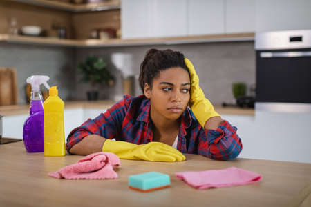 Sad tired despaired millennial black housewife in rubber gloves holding her head with handの写真素材