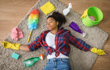 Tired millennial african american woman housewife lying down on floor with cleaning supplies in room...