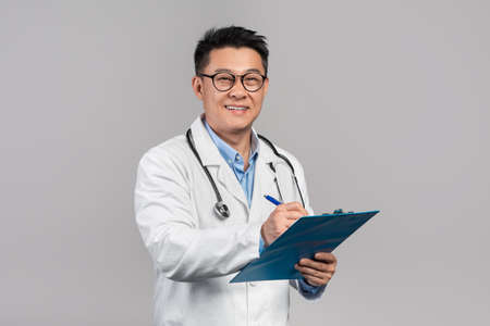 Cheerful mature korean man doctor in white coat, glasses with stethoscope writes at tablet