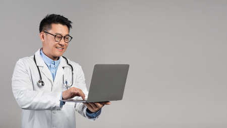 Glad smiling confident middle aged asian doctor in white coat, glasses with stethoscope typing on laptopの写真素材