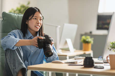 Cheerful Vietnamese Woman Photographer Holding Photocamera Sitting At Workplaceの写真素材