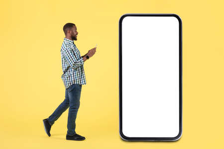 Black Male Using Smartphone Walking Near Huge Phone, Yellow Background