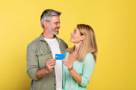 We love our bank. Happy caucasian mature spouses with credit card looking at each other and smiling,...