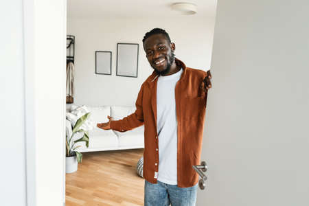 Welcome. Happy african american man inviting visitor to enter his home, standing in doorway of modern apartmentの写真素材