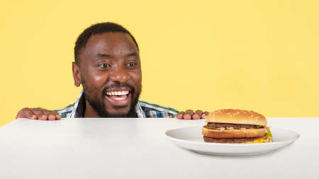 African Guy Looking At Burger On Table Over Yellow Backgroundの写真素材