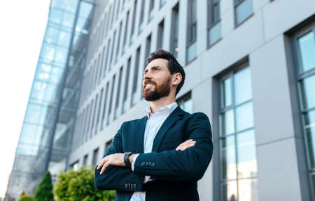 Smiling confident handsome young businessman with beard in suit with crossed arms on his chest looks at empty spaceの写真素材