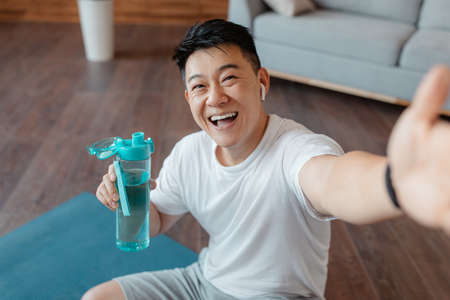 Happy mature asian man with bottle of water taking selfie on break from home workout, sitting on fitness matの写真素材