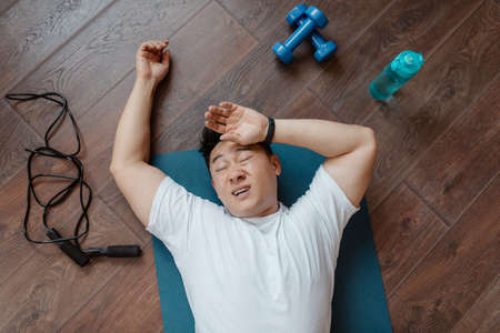 Fatigue after exercising at home. Tired mature asian man wiping sweat, lying on mat on floor with...