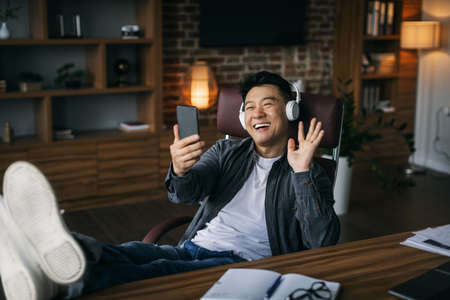 Happy middle aged asian male in headphones rest, has video call on phone, waving hand in home officeの写真素材
