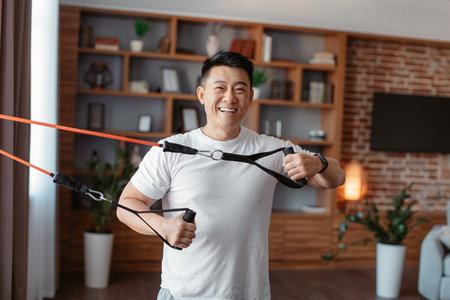 Home fitness. Happy asian man exercising using resistance rubber band, training in living room at...
