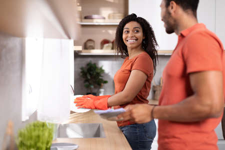 Smiling young african american husband and wife in same t-shirts washing plates together in kitchen interiorの写真素材