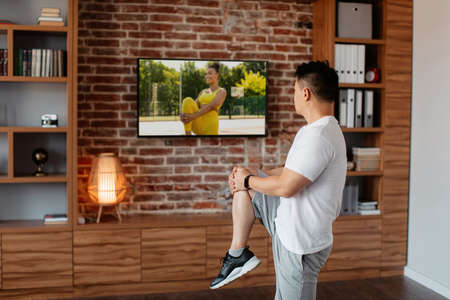 Active asian mature man exercising at home and watching fitness workout program on TV, doing warm up exercisesの写真素材