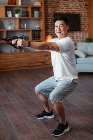Excited active asian man doing sport workout in living room, exercising with resistance band and...