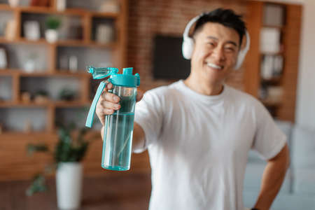 Smiling active asian middle aged man in sportswear wearing headphones and holding bottle of water,...