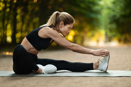 Sporty blonde woman stretching on yoga mat at parkの写真素材