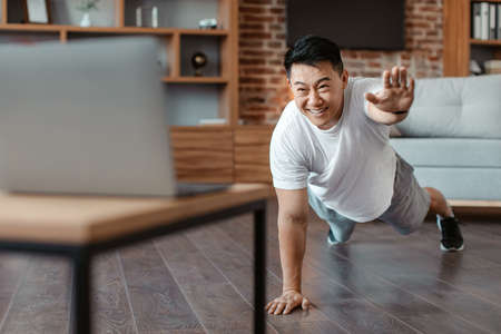 Online sports. Sporty asian mature man doing exercises near laptop, standing in plank pose and raising one handの写真素材