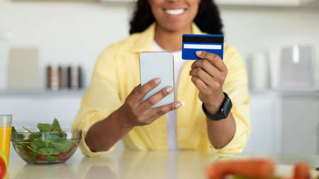 Grocery delivery app. Young black woman using cellphone and credit card, ordering food and groceries productsの写真素材