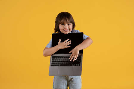 Modern technologies and kids. Cute little boy carrying open laptop and smiling to camera, yellow studio backgroundの写真素材