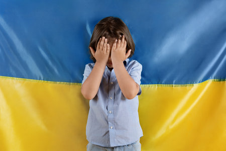 Stop killing kids at war. Little scared boy crying, hiding his face with hands, posing over Ukrainian flagの写真素材