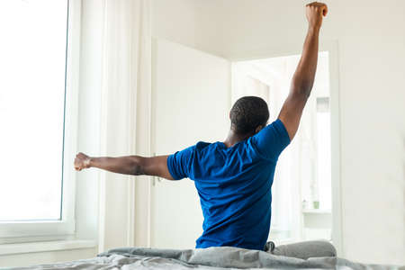 Black Male Stretching Arms Sitting Back To Camera In Bedroomの写真素材