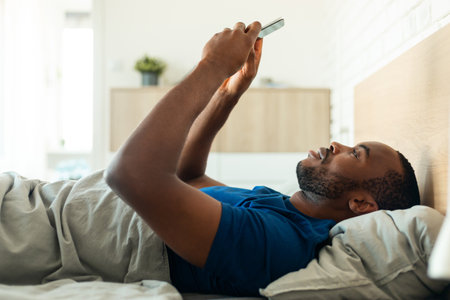 African American Guy Using Phone Lying On Back In Bedroomの写真素材