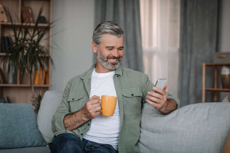 Glad retired european man with beard chatting on smartphone, enjoying drink and chat in social...