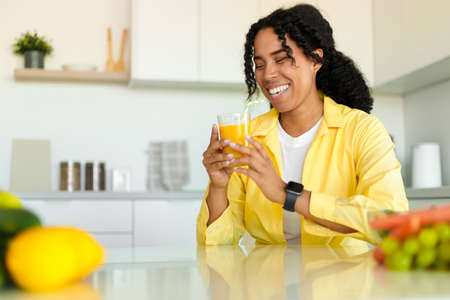 Excited black lady drinking fresh orange juice at home, sitting at table in kitchen interior and...