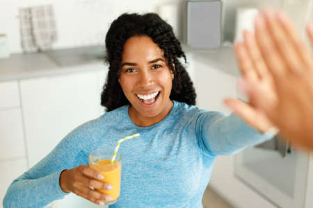 Happy african american fit lady drinking fresh juice and giving high five to friend, standing in...