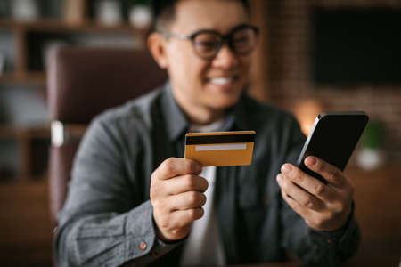 Cheerful adult japanese man in glasses typing on phone, show credit card, shopping online, invested money to startupの写真素材