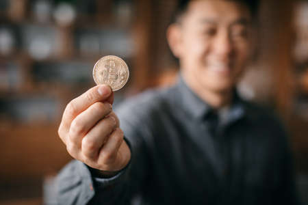 Smiling successful adult chinese man show bitcoin coin, manager work with cryptocurrency in office...