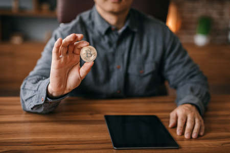 Mature man show bitcoin coin at table with tablet with blank screen in office interior, cropped, close upの写真素材