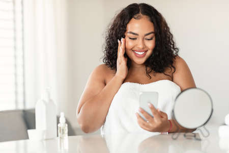 Attractive black bodypositive woman in white towel sitting in bedroom, touching her face and using smartphoneの写真素材