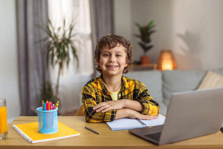 Great online classes. Cute boy sitting at desk and smiling to camera, enjoying homeschooling on...