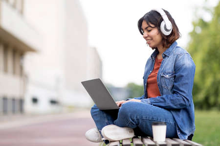 Smiling arab female student in wireless headphones study with laptop outdoorsの写真素材