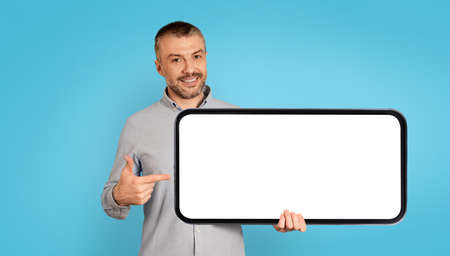 Cheerful Man Holding Large Smartphone With Empty Screen, Blue Backgroundの写真素材
